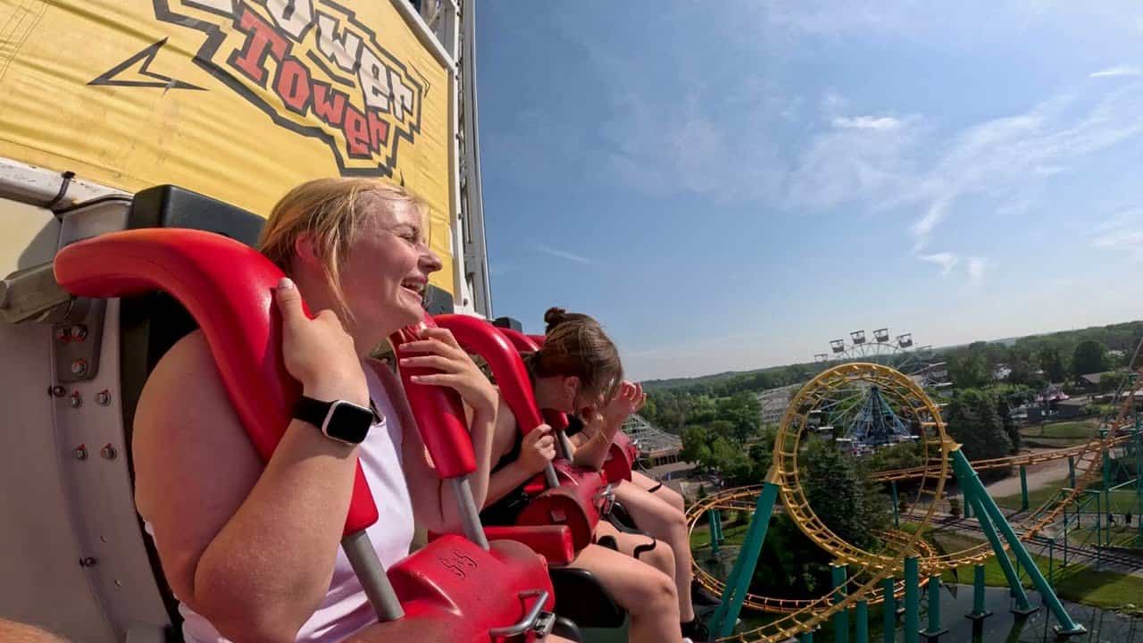 Two young women are seated in a red and white amusement park ride with their harnesses secured, one laughing and the other covering her face. The "Power Tower" sign looms above them. In the background of the Six Flags theme park, a clear sky, green landscape, and roller coasters complete the thrilling scene at Valleyfair theme park. 