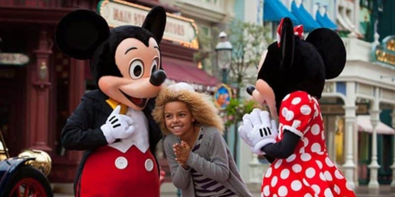 A young girl with curly hair stands excitedly between two costumed characters, Mickey Mouse and Minnie Mouse, on a colorful street. Mickey wears his classic red pants and Minnie is dressed in her signature red polka dot dress.