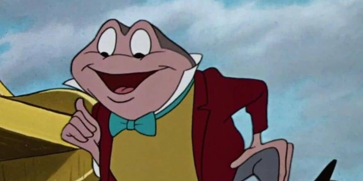 An illustrated amphibian character in a red coat, yellow vest, and blue bow tie, smiling and gesturing with his right hand. The background includes a partially visible yellow structure and a cloudy sky, reminiscent of scenes from the Magic Kingdom.