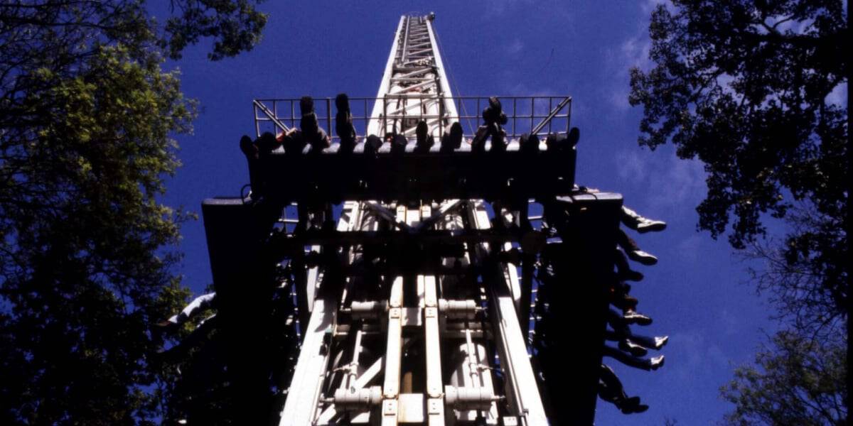 A low-angle view of a theme park ride captured against a bright blue sky. Riders are seated and dangling their legs as they prepare for an exhilarating drop. Tall trees frame both sides of the image, accentuating the height of the ride.