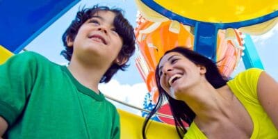 A young boy in a green shirt and a woman in a yellow shirt, both smiling and looking up, are enjoying a sunny day at an amusement park. They appear to be inside a colorful theme park ride, with blue, yellow, and orange elements around them.