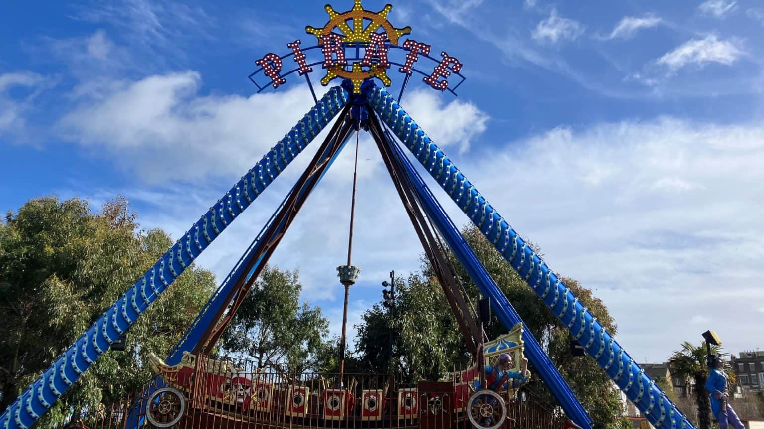 A large pirate ship-themed amusement ride swings back and forth against a backdrop of trees and a blue sky with scattered clouds. The word "Pirate" is displayed in lights at the top of the ride. The ride is surrounded by gates adorned with nautical decorations.