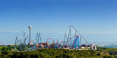 A panoramic view of PortAventura theme park with various roller coasters featuring blue and red tracks, surrounded by greenery. A tall observation tower stands on the left, with clear blue skies and distant mountains in the background, offering visitors free entrance.