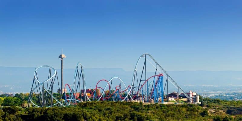 A panoramic view of PortAventura theme park with various roller coasters featuring blue and red tracks, surrounded by greenery. A tall observation tower stands on the left, with clear blue skies and distant mountains in the background, offering visitors free entrance.