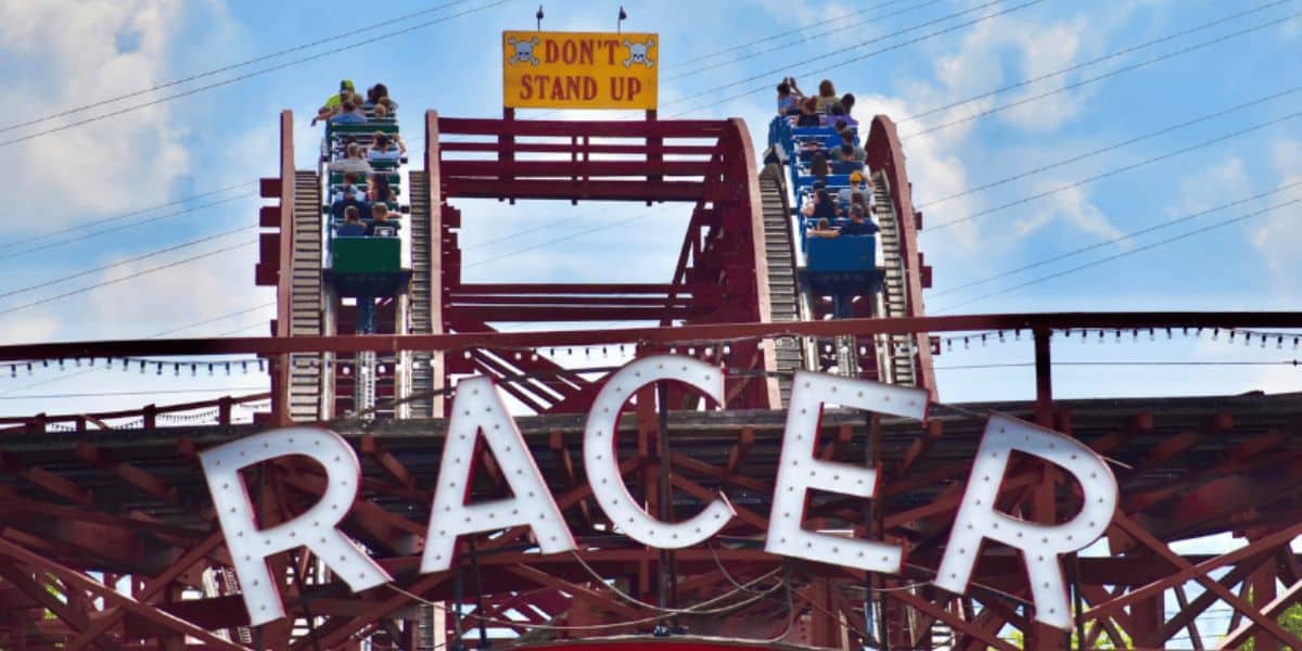 A theme park roller coaster named "Racer" features two parallel tracks with riders approaching a drop. A sign at the top reads "DON'T STAND UP." The 97-year-old roller coaster rides under a clear sky with a few clouds visible.