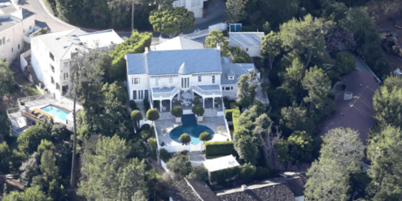An aerial view of a large, white, two-story house surrounded by trees and greenery. The property, perfect for an LA fitness guru or entertainer, features a well-maintained garden, a swimming pool, and a patio area with outdoor seating. The house is in a residential neighborhood with other homes nearby.