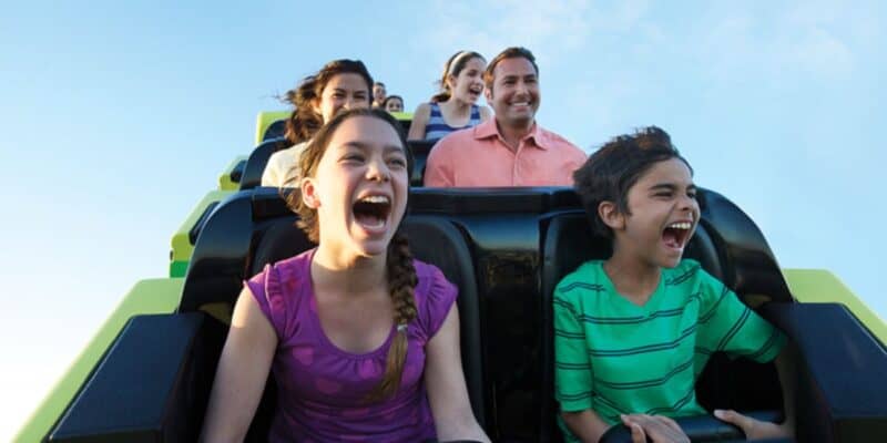 A group of people riding a roller coaster at SeaWorld are depicted in the image. The front row consists of two excited kids, one wearing a purple shirt and the other in a green striped shirt, both screaming with joy. In the background, the rest of the riders share similar expressions of excitement.