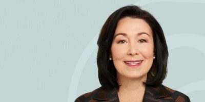 A woman with shoulder-length dark hair, wearing a brown patterned jacket and pearl earrings, smiles at the camera. The background is light blue with subtle, abstract white oval shapes.