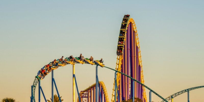 A vibrant roller coaster at the theme park SeaWorld features yellow, purple, and blue tracks twisting against a clear sky at sunset. Filled with passengers, some with arms raised, they experience the thrill and challenge of a steep climb and sharp turns. Palm trees are visible in the background.