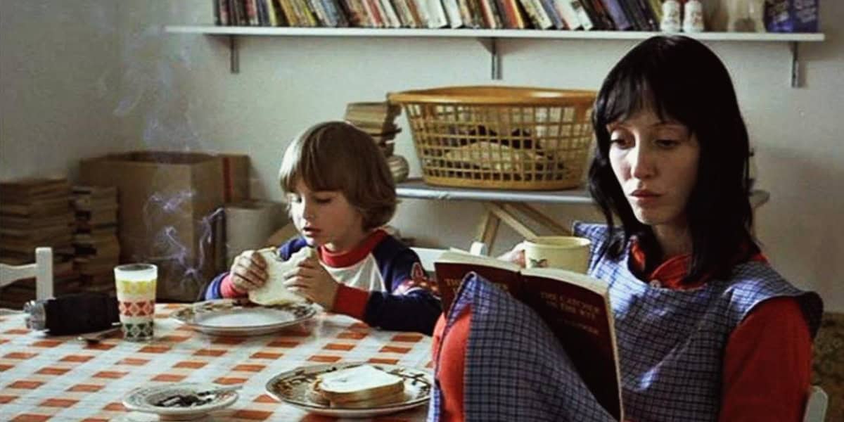 In a cozy kitchen with a checkered tablecloth, a woman with dark hair reads a book while holding a cup, and a young child with light hair and a red and blue shirt eats at the table. A steaming mug and some bread are on the table. Shelves with books, including one on legendary horror icons, are in the background.
