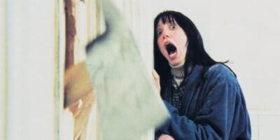 A woman with long dark hair and wide eyes screams in terror as a blurred axe breaks through a door beside her. In her blue sweater over a turtleneck, she embodies the fear that legendary horror icons are famous for. The white background makes the scene even more dramatic and tense.