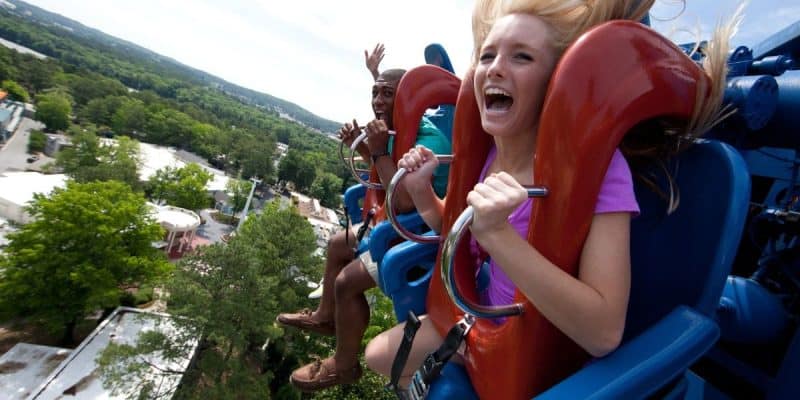 Two people, a woman in a pink shirt in the foreground and a man in a green shirt in the background, are enjoying a roller coaster ride at Six Flags. Both are smiling with their hands in the air. The ride is high above green trees and a park area in the background.