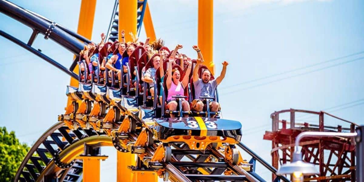 A group of people are riding a roller coaster with their hands in the air, expressing excitement and thrill. The vibrant roller coaster features bright yellow supports and a loop in the background under a clear blue sky.