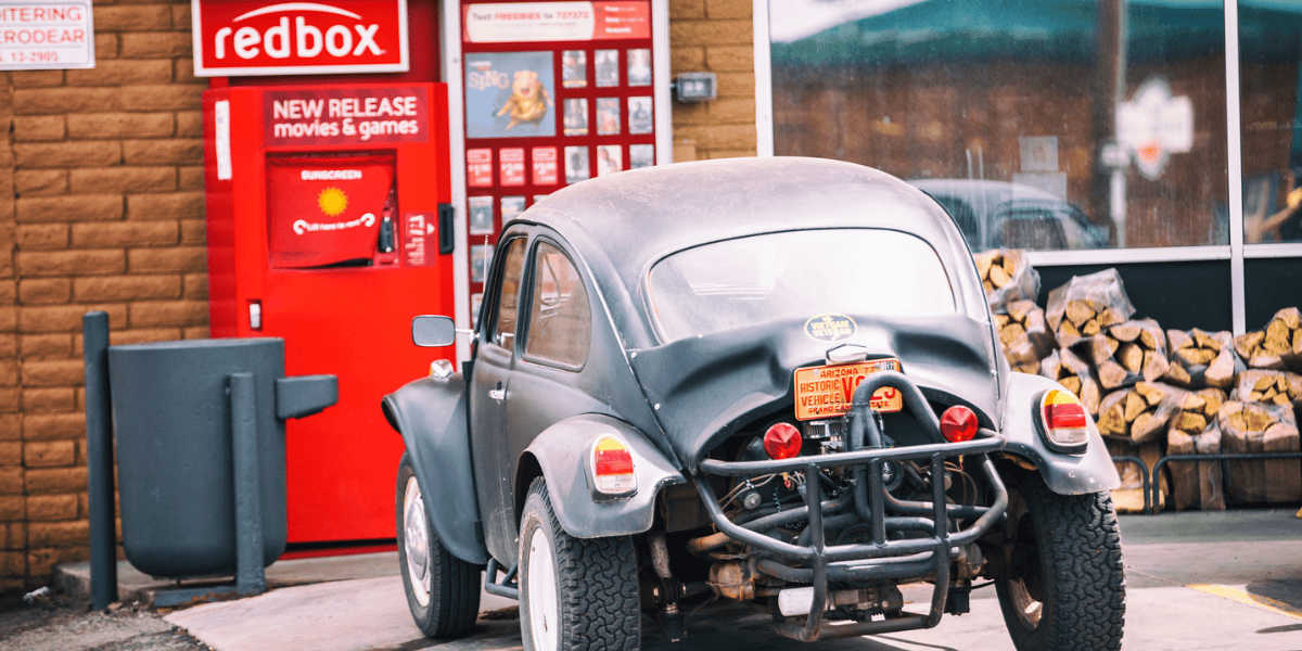 A black vintage car with a rugged exterior is parked in front of a Redbox kiosk at a convenience store. Stacked firewood is visible on the right, and posters for new movies from a popular streaming service are displayed on the kiosk.