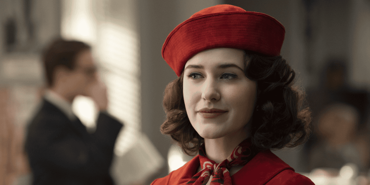 A woman with wavy brown hair is smiling slightly, wearing a red hat and a matching scarf. She is dressed in a red coat. The background shows a blurred figure of a man in a suit, holding a book with his hand raised to his chin. It looks like they're discussing the recent Chapter 11 Bankruptcy filing indoors.