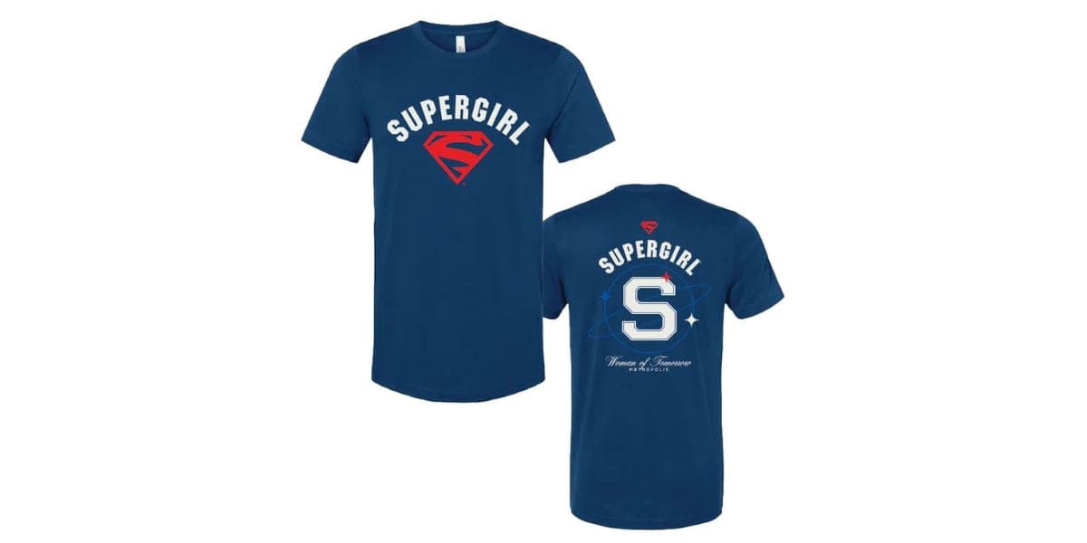 Front and back view of a blue t-shirt with "SUPERGIRL" written in white at the top. The front features a red Supergirl logo, while the back showcases a large letter 'S' with "SUPERGIRL: Woman of Tomorrow" and small star patterns.