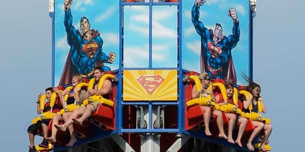 People are enjoying an amusement park ride themed after Superman at Six Flags. The ride features two large illustrations of Superman in flight on a blue background. The riders appear thrilled, with some raising their hands and others with hair flying.