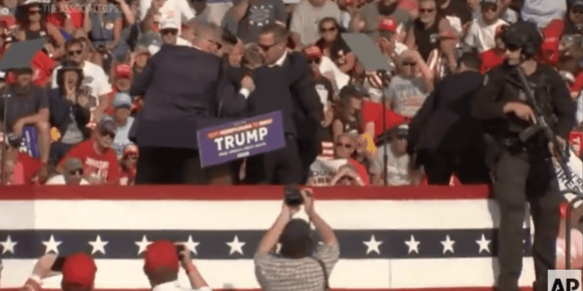 Security personnel react to an incident during a crowded outdoor rally with a "Trump" sign visible on the podium. Amid rally chaos, a large audience, many wearing red hats, watches the scene while a photographer captures the moment in the foreground.