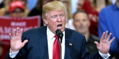 Donald Trump wearing a black suit, white shirt, and red tie, is speaking into a microphone. His hands are raised, and his mouth is open in mid-speech. A crowd is visible in the background, along with a sign that partially reads "MAKE AMERICA," reminiscent of a scene from Captain America 4 within the MCU.