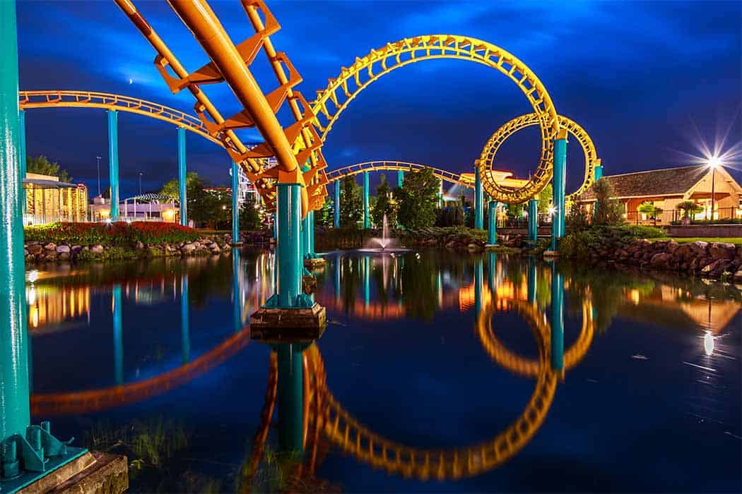 A vibrant, twisting roller coaster with yellow tracks and blue supports at Six Flags is illuminated at night. The coaster is reflected in a calm pond below, creating a mirror image. Lush greenery, buildings, and a water fountain are visible in the background of the theme park scene at Valleyfair theme park.