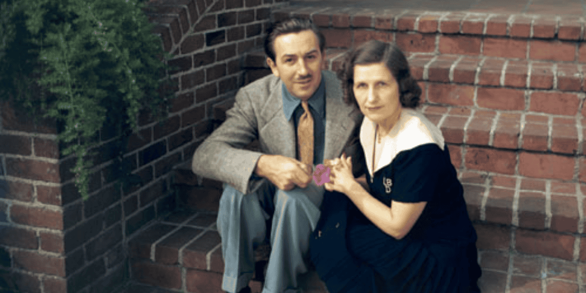 A man and a woman sit close together on a set of brick stairs, holding hands. The man is wearing a gray suit, light blue shirt, and yellow tie. The woman is in a dark dress with a white collar. Both are looking at the camera. A leafy plant is in the background.