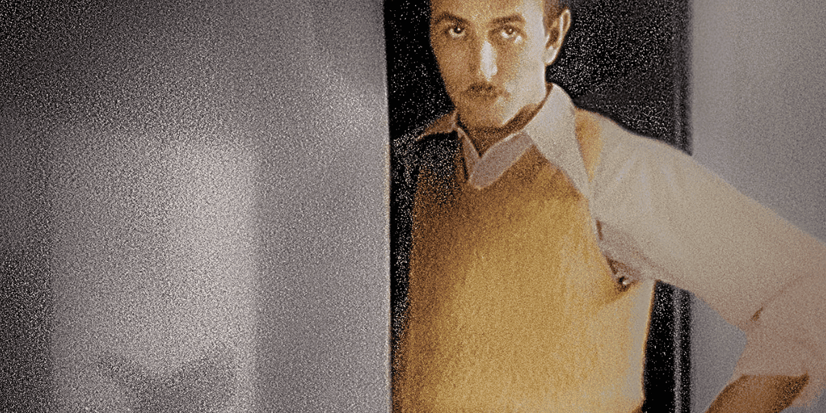 A person with short hair, wearing a yellow vest over a long-sleeve shirt, stands with one arm leaning on a doorframe. The background is out of focus, and the image has a grainy texture. The person has a serious expression and looks towards the camera.