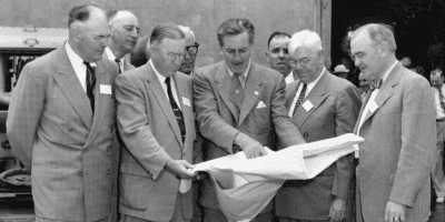 A group of six men in suits stand closely together, looking and pointing at a large unfolded paper one man is holding. They appear to be discussing its contents. The background shows an outdoor setting with a vehicle and other people in the distance.