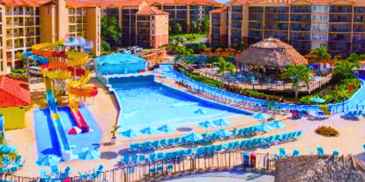 A vibrant resort features multiple pools, water slides, and a splash playground. There are numerous lounge chairs with umbrellas around the pools. This Disney World competitor includes multi-story buildings in the background and a thatched-roof hut area surrounded by palm trees.
