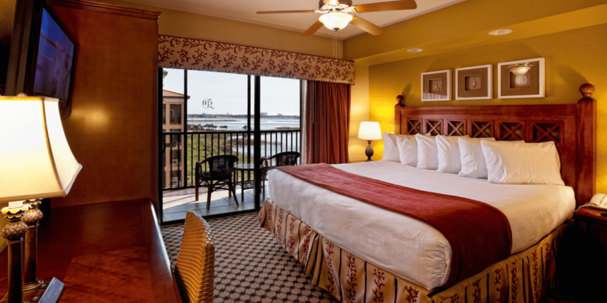 A warmly lit hotel room with a king-sized bed adorned with white pillows and a red runner. A ceiling fan is above, and a desk with a lamp and TV is on the left. Sliding doors open to a balcony with outdoor seating, overlooking a serene lake view—a perfect retreat from exploring Disney World Competitor parks.