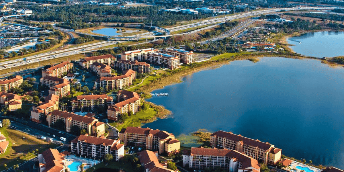 Aerial view of a large Disney World competitor resort with multiple orange-roofed buildings near a calm lake. Surrounding the resort are green areas and roads, with a major highway in the background. Small patches of water and greenery are also visible in the distance.