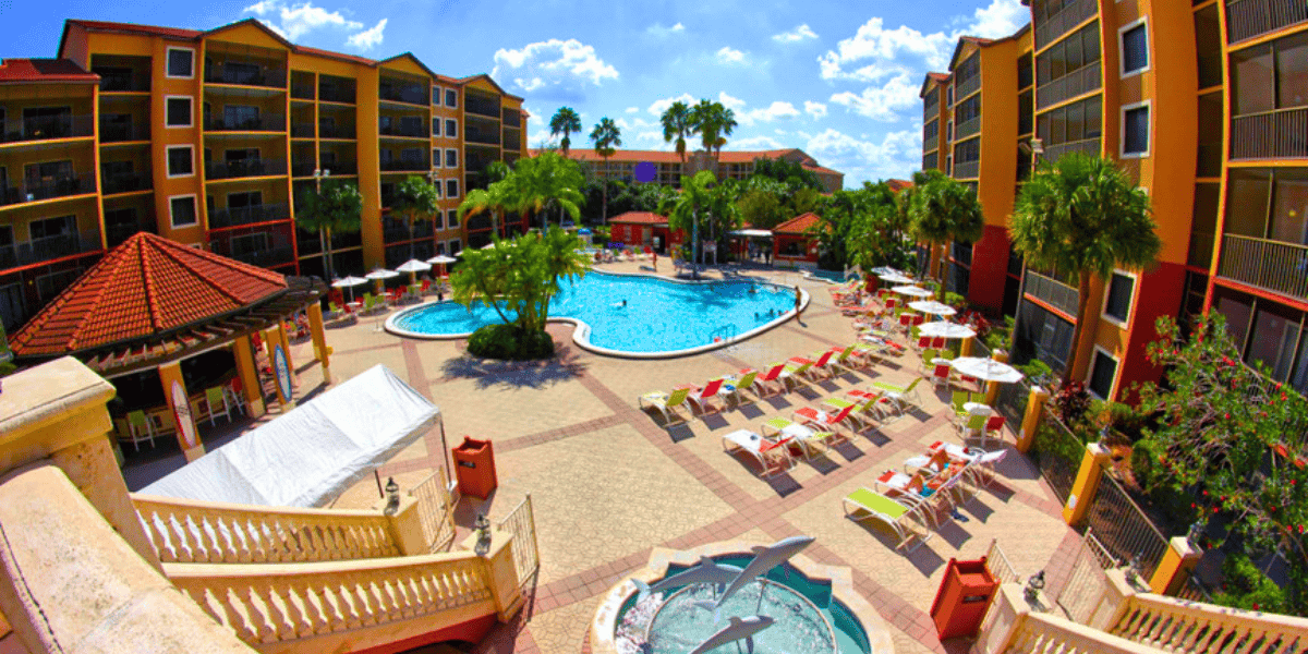 A bright, colorful resort courtyard with a large circular swimming pool surrounded by lounge chairs and umbrellas. Palm trees and multi-story buildings with balconies encircle the central area. A smaller circular kiddie pool with a dolphin fountain is in the foreground, reminiscent of Disney World magic.