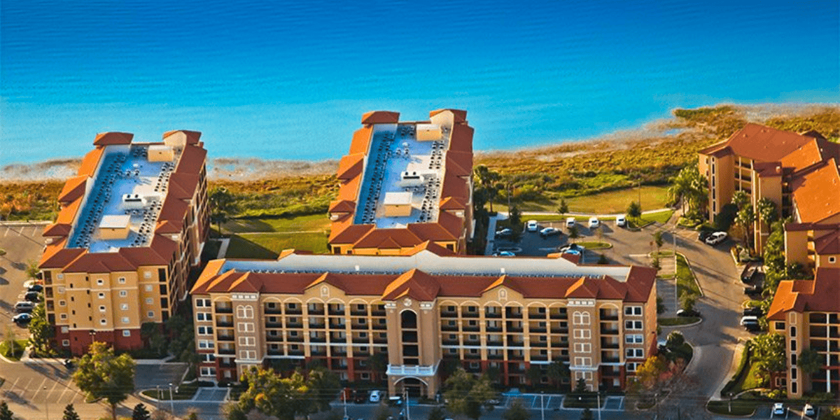 Aerial view of a waterfront resort, Disney World competitor, featuring three large buildings with red roofs and balconies, surrounded by lush greenery and parking areas. The vibrant blue water and shoreline are visible in the background, creating a serene and picturesque setting.