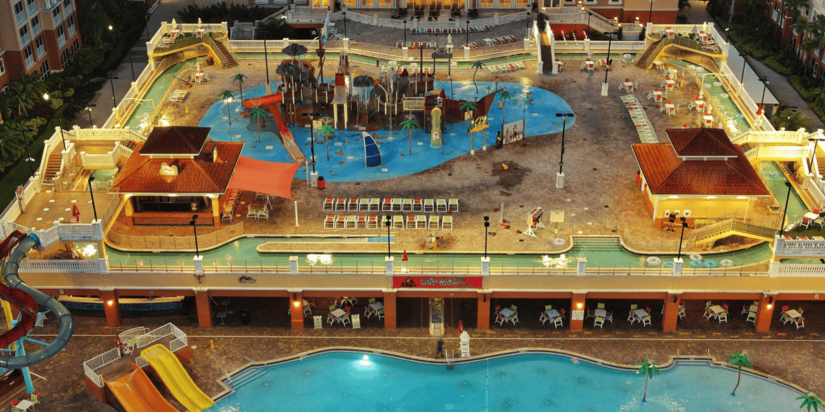 Aerial view of an extensive outdoor water park at dusk. Features multiple water slides, lounging areas with colorful chairs, and a lazy river encircling the park. The pool area in the foreground glows under lighting, with resort buildings visible in the background—a true Disney World competitor.