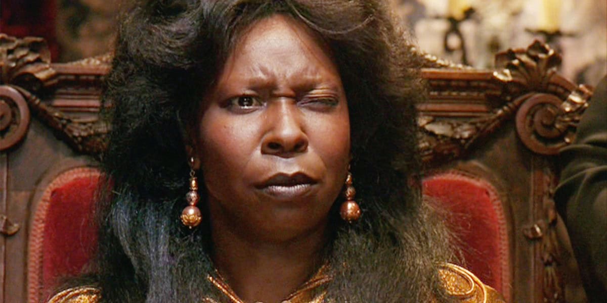 A person with long, dark hair and wearing dangling earrings is sitting on an ornate chair. They are winking one eye while the other is open wide, and they have a concerned or confused expression on their face. The background is blurred and appears to be indoors.