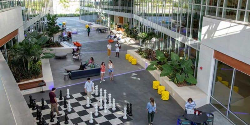 An outdoor courtyard of Disney’s Flamingo Crossings features a large chess set with human-sized pieces. People are engaged in various activities, such as playing chess, walking, and sitting on benches. The area is decorated with plants, and yellow seating cubes are placed around, enhancing the chance to live at Disney.