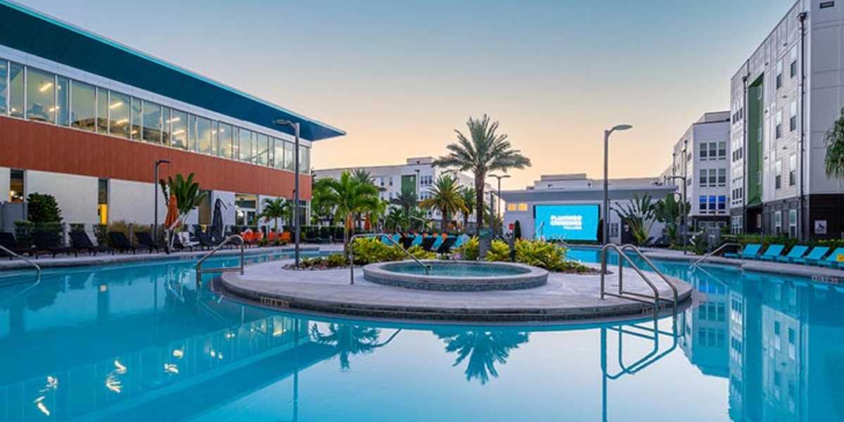 A modern resort pool area at dusk features a large, multi-leveled swimming pool surrounded by palm trees and lounge chairs. Nearby, Disney’s Flamingo Crossings and a well-lit, multi-story structure are visible in the background, reflecting the vibrant ambiance and offering guests a chance to live in luxury.