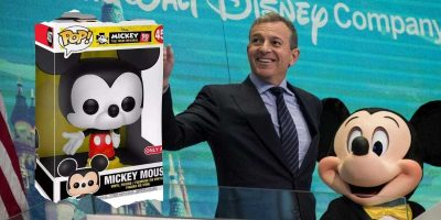 A man in a suit smiles and waves while standing next to a person in a Mickey Mouse costume. To their left, there is a large Mickey Mouse Funko Pop! figurine in its box. The background shows the Disney logo and a castle image, possibly hinting at an exciting Toy Venture overseen by the Disney CEO.