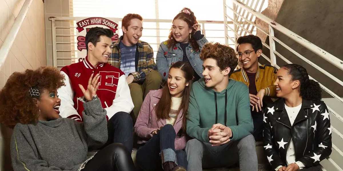 A group of eight young adults sits on a staircase, smiling and chatting. They could easily be mistaken for the cast of "High School Musical," with some wearing casual clothes and others in school attire. Behind them, a sign reads "East High School." The setting looks like a school hallway.