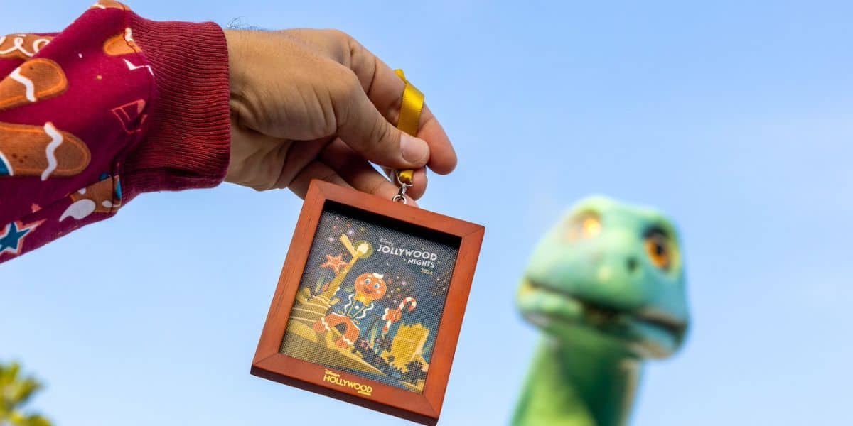 A person wearing a Jollywood Nights spirit jersey with Christmas-themed designs holds a framed Jollywood Nights holiday decoration ith Ollie, perfect for Disney Jollywood Nights Merchandise. In the background, a large green dinosaur statue is slightly blurred against a clear blue sky, capturing the festive Holiday Season.
