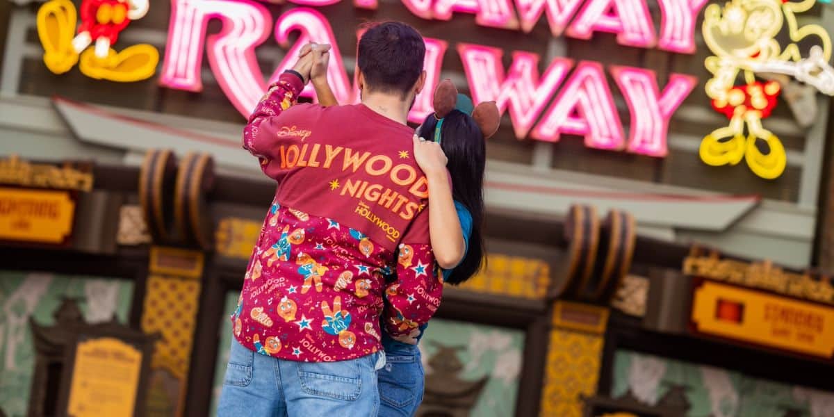 A couple stands close together with their backs to the camera, holding hands and gazing at a neon sign that reads "Runaway Railway." The man is wearing a red Jollywood Nights spirit jersey with Disney characters, and the woman has Minnie Mouse ears on. It’s the holiday season, and they’re at Disney World celebrating Disney Jollywood Nights.