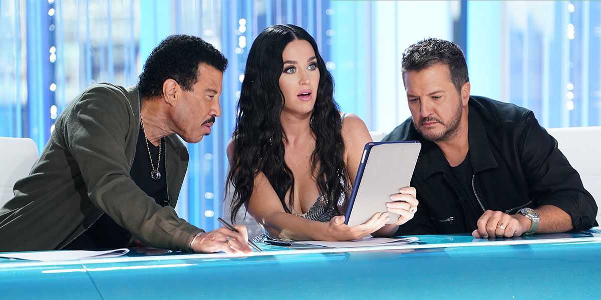 Three people are seated at a desk, examining a piece of paper or tablet in front of them. The person in the middle appears surprised or intrigued, the person on the left is closely inspecting the paper, and the person on the right is looking attentively.