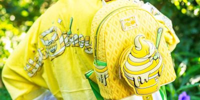 A yellow backpack with a cartoon ice cream design is worn by a person. The backpack features a Pineapple Swirl soft serve cone image with yellow accents and green details. The person, donning a yellow-themed outfit with cartoon-like imagery, could be straight out of Walt Disney World's new collection. The background is blurred greenery.
