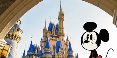 An animated version of Mickey Mouse is seen on the right side of the image, with a slightly sad expression. In the background, there's a view of Cinderella's Castle at Disney World, framed by an archway and featuring distinct blue and gold spires. A scooter user can be seen enjoying their visit to the park.