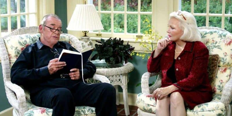 An elderly man, wearing glasses and dressed in a black outfit, reads a book while seated on a floral upholstered chair. Sitting across from him on a similar chair, an elderly woman with white hair, dressed in a red outfit, reminiscent of Gena Rowlands in *The Notebook*, listens attentively and holds a pen to her lips.