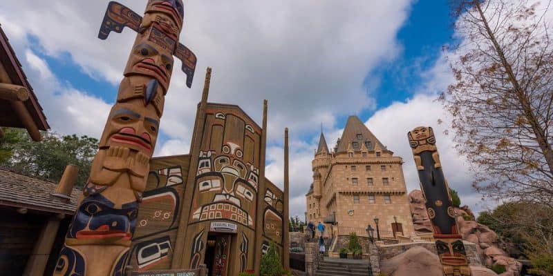 The image shows a picturesque area featuring tall wooden totem poles and intricate carvings on a wooden structure, reminiscent of a forgotten park. In the background, there's a castle-like stone building under a partly cloudy sky, with trees flanking the sides. A few people are seen walking in the distance.