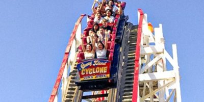 A group of excited people, some with their hands raised, are enjoying a ride on the Coney Island Cyclone roller coaster. The ride is at the peak of a climb, with a clear blue sky in the background. The front car of the coaster displays "Coney Island Cyclone" signage.