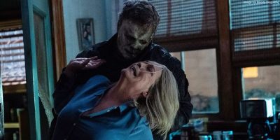 A masked figure dressed in dark clothing grabs an older woman by the neck from behind in a dimly lit room. The woman, wearing a blue shirt, appears distressed and is looking upward.