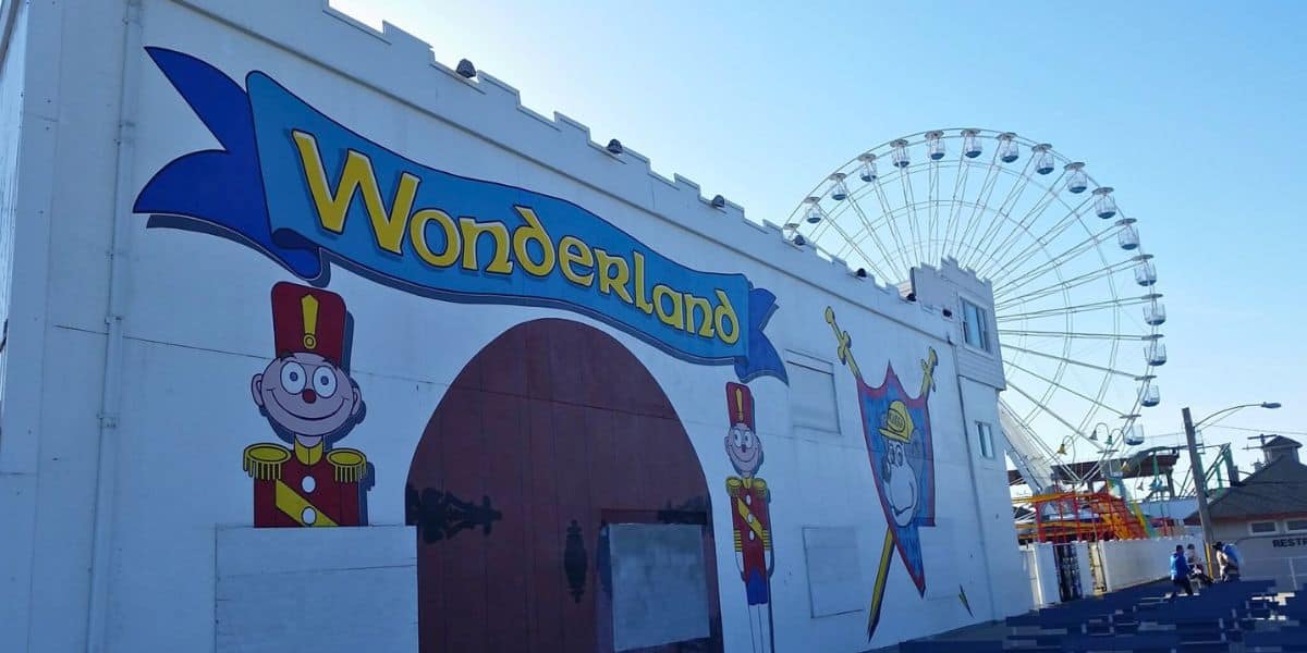 A funfair scene features a white building with the word "Wonderland" in bold, colorful letters and cartoon soldier characters painted on the wall. Behind the building, a large Ferris wheel rises into the clear blue sky. This historic theme park has delighted visitors for over 100 years, titled Gillian's Wonderland Pier.