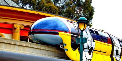 A sleek, colorful monorail featuring a large painted image of a Disney cartoon character's face glides along an elevated track. The station in the background has classical architectural elements and is surrounded by tall trees. A vintage-style lamppost is in the foreground, symbolizing timeless monorail service.