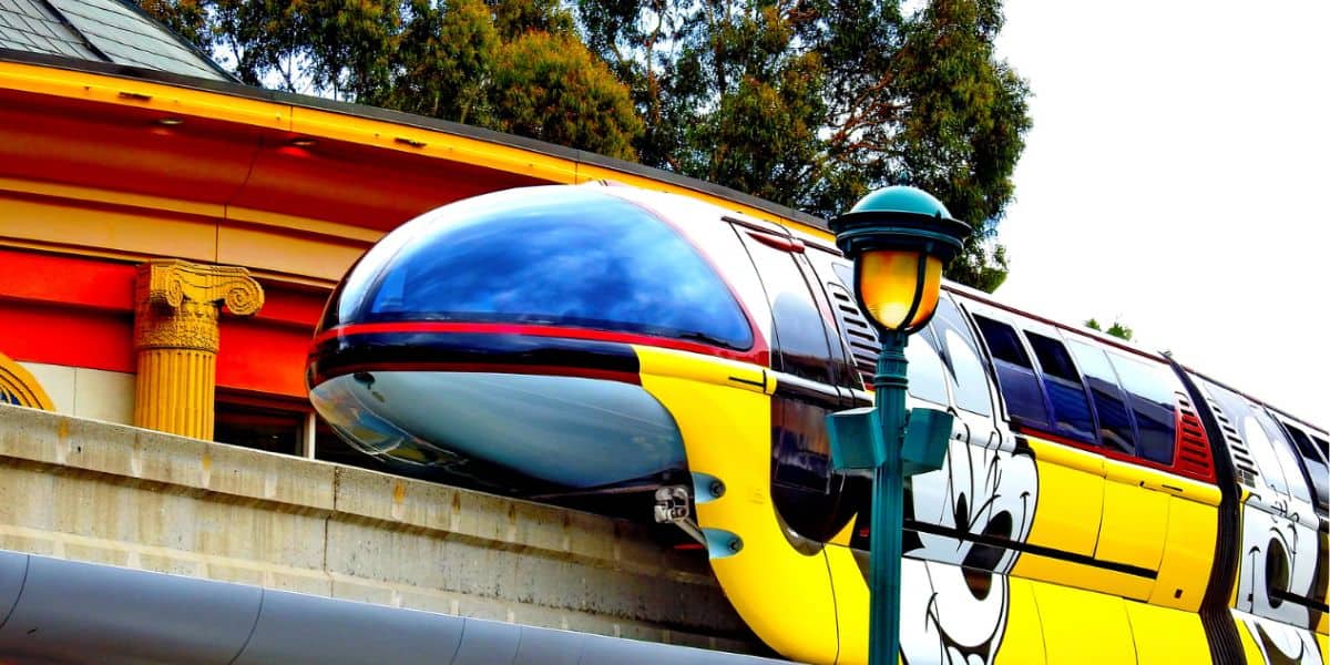 A sleek, colorful monorail featuring a large painted image of a Disney cartoon character's face glides along an elevated track. The station in the background has classical architectural elements and is surrounded by tall trees. A vintage-style lamppost is in the foreground, symbolizing timeless monorail service.
